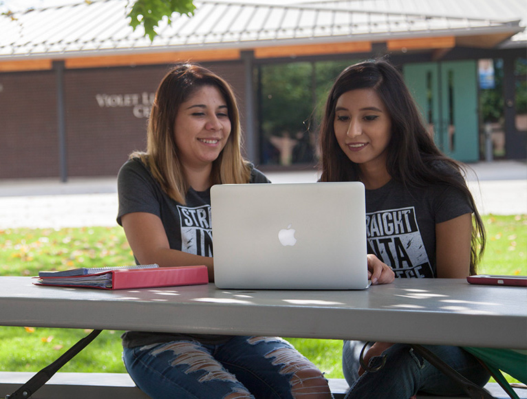 Two Female Students Looking At Computer Outside On Campus