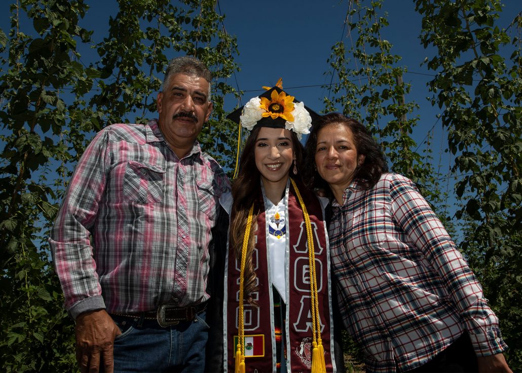 Maria (dressed in graduation regalia) and her parents pose in a hop field