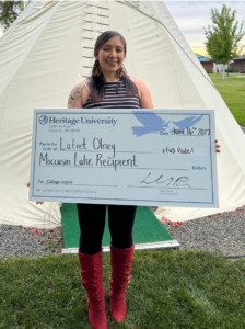 Young woman in front of a Native American tepee holding a large check representing a scholarship award.