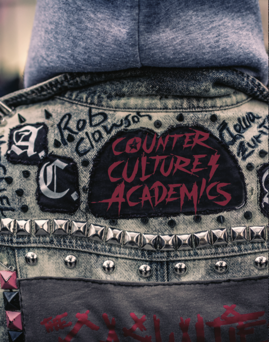 Closeup of a denim jacket decorated with metal studs, letters in a fancy font with the words Counter Culture Academics written in red on a black patch