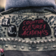 Closeup of a denim jacket decorated with metal studs, letters in a fancy font with the words Counter Culture Academics written in red on a black patch