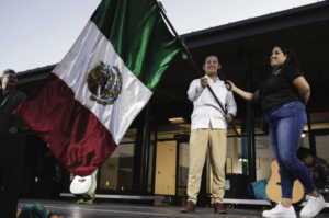 A man holds the Mexican flag as a woman holds a microphone to his face