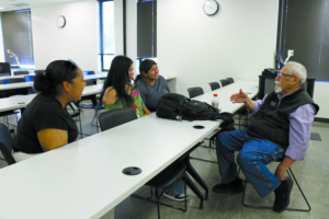 A professor sitting at a table across from three students in a classroom.