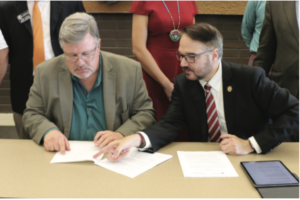 Two men sit at a desk signing papers while people stand behind them.