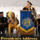 People in Native American regalia on a stage with people in higher education regalia paying attention to the university president's address