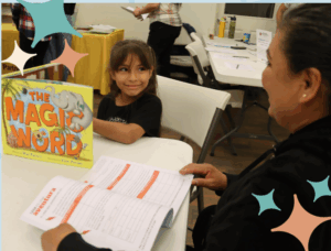 A mother and her child sit at a table while her child who's holding a book smiles at her
