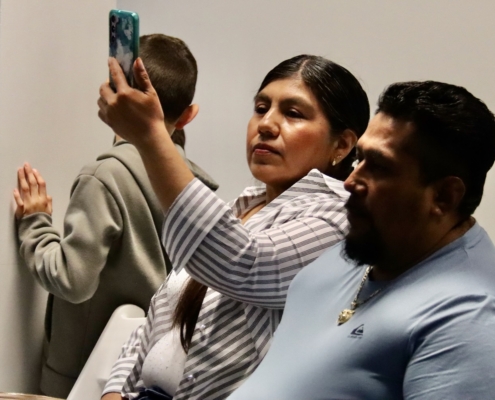 Photograph showing three people sitting closely together, with one person holding up a smartphone to take a selfie or photo. The individuals wear casual clothing, including a striped shirt and a light blue shirt, and are positioned against a plain background.