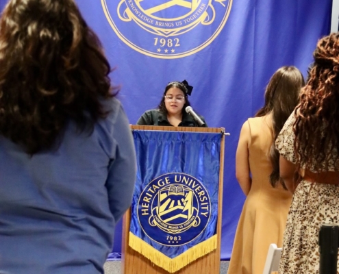Photograph of a speaker standing at a podium with Heritage University banners and a large emblem displayed on a blue backdrop behind. Audience members stand facing the speaker, with the university's name, founding year 1982, and logo prominently featured on both podium and backdrop.
