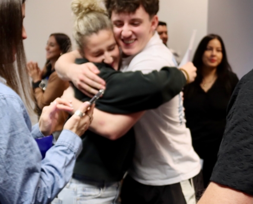 Photograph showing a group of people in a social setting, with two individuals embracing in the center. The scene includes casual clothing, blurred faces, and others engaged with mobile devices, suggesting a moment of connection or reunion.