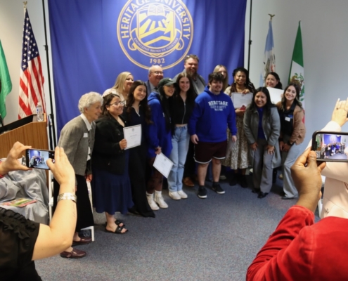 Photograph of a group of people posing for a photo in front of a blue Heritage University banner with flags on either side. Several individuals hold certificates, while others take pictures with smartphones, indicating a celebratory or award event.