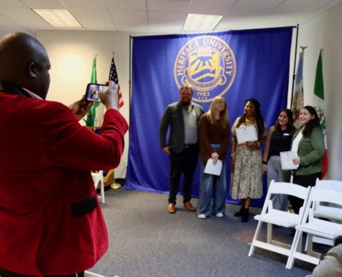 Photograph of a group photo session at Heritage University, showing four people standing in front of a blue backdrop with the university seal, while a man in a red jacket takes their picture with a smartphone. The setting includes American and Mexican flags, white folding chairs, and individuals holding certificates, indicating a celebratory or award event.
