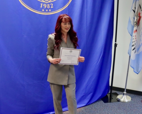 Photograph of a person standing in front of a blue backdrop with a gold emblem and text reading "Knowledge Brings Us Together 1982," holding a certificate. The setting includes a flag on a stand to the right, suggesting a formal or official recognition event.