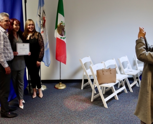 Photograph of a small group posing for a photo in front of blue and white flags, including a Mexican flag, in an indoor setting. Four people stand together on the left, one holding a certificate, while a fifth person on the right takes their picture with a smartphone; several empty white folding chairs and a handbag are visible in the background.