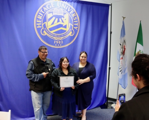 Photograph of three people standing in front of a blue backdrop with Heritage University logo and flags on the right side, with one person holding a certificate. Another person is taking a photo of the group using a smartphone, capturing a moment of recognition or achievement.