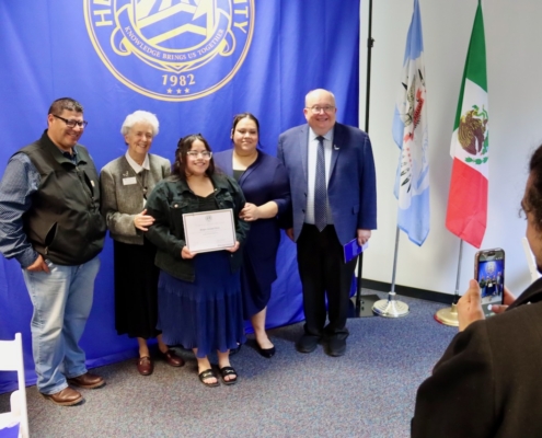Photograph of a group of five people standing together in front of a blue backdrop with a university emblem and flags of Mexico and another organization. One person in the center holds a certificate while another individual on the right takes a photo with a smartphone, indicating a formal recognition or award ceremony.