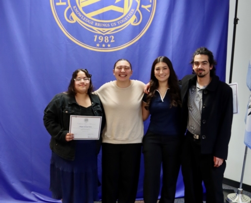 Photograph of four people standing in front of a blue Heritage University backdrop with the university logo and founding year 1982. One person holds a certificate, indicating a recognition or graduation event, with all individuals dressed in semi-formal attire.