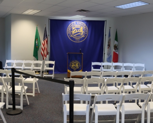 Photograph of a small event setup at Heritage University featuring rows of white folding chairs facing a podium with a microphone, positioned in front of a blue backdrop displaying the university seal. Flags of the United States, Washington state, Illinois, and Mexico flank the backdrop, indicating a formal or multicultural gathering space.