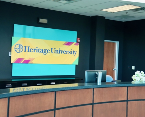 Photograph of a reception area featuring a curved wooden desk with a black countertop and a bouquet of white flowers on the right side. Behind the desk, a bright yellow and blue sign displays "Heritage University" with a logo, set against a teal wall with a door and dark walls in the background.