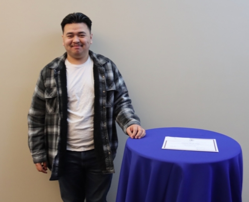 Photograph of a person standing next to a round table covered with a blue tablecloth, featuring a single document placed on top. The person is wearing a black and white plaid jacket over a white shirt and dark pants, positioned against a plain beige background.