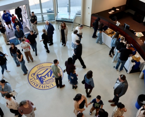 Photograph showing a university lobby with people gathered around food tables and socializing. The floor features a large university emblem in blue and gold, indicating a campus event or reception.