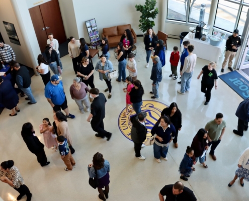 Photograph showing a diverse group of people gathered in a spacious indoor area with a university logo on the floor. The scene captures social interaction and movement, with individuals of various ages and attire standing, walking, and conversing near seating and entrance areas.