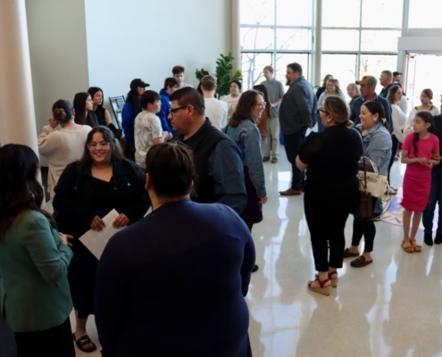 Photograph of a large group of people gathered indoors, engaged in conversations and waiting in line near a registration or information desk. The setting features bright natural light from large windows and a polished floor, indicating a formal or organized event such as a conference or seminar.