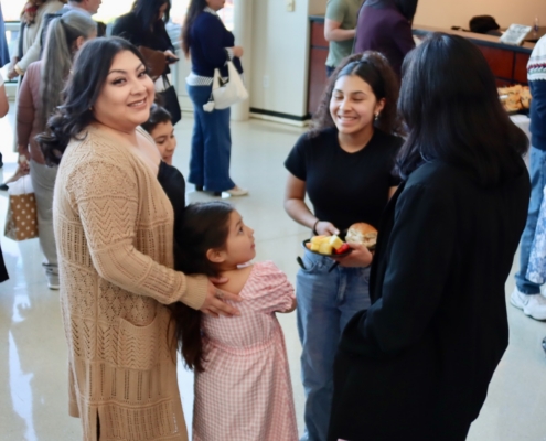 Photograph showing a group of people gathered indoors, some standing in line while others engage in conversation. Key details include a woman in a beige cardigan holding a young girl in a pink dress, and another person holding food items, indicating a social or community event with casual interaction.