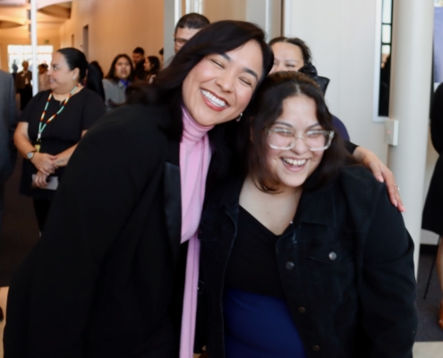Photograph of two people posing together in a crowded indoor setting, likely at a social or professional event. Background shows several other individuals standing and interacting, with warm lighting and neutral-colored walls.