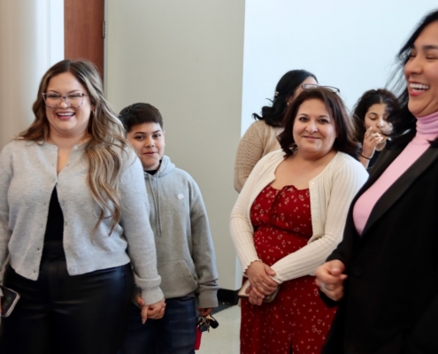 Photograph of a group of people standing indoors, engaged in conversation or listening attentively. Individuals wear casual to semi-formal clothing, including a red dress with white cardigan, gray sweaters, and a black blazer, suggesting a social or professional gathering.