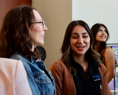 Photograph showing a group of people engaged in conversation indoors, with one person wearing a black shirt and brown jacket featuring a logo. The setting includes a wooden pillar and a blurred background with a screen displaying colorful content.
