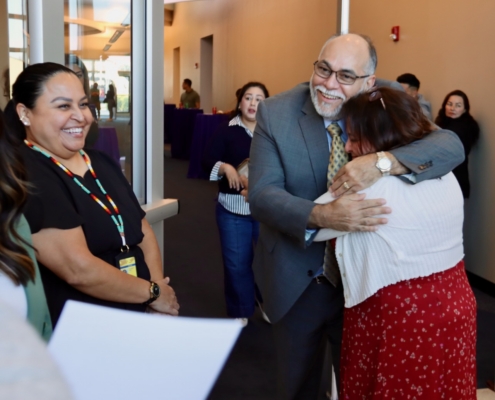 Photograph showing a group of people indoors, with one man in a suit and tie embracing a woman in a red skirt and white top. Other individuals stand nearby, some holding papers, suggesting a formal or emotional gathering, possibly a meeting or event.