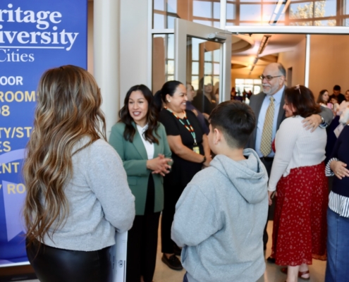 Photograph of a group of people gathered in a hallway near a blue sign for Heritage University Tri-Cities, indicating classroom and office locations. The setting appears to be an event or orientation, with individuals engaged in conversation and wearing casual to business-casual attire.