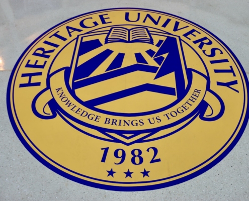 Seal of Heritage University displayed on a floor, featuring a circular design with blue and gold colors. The seal includes an open book, mountains, and rays, with text "Knowledge Brings Us Together," founding year "1982," and three stars at the bottom.