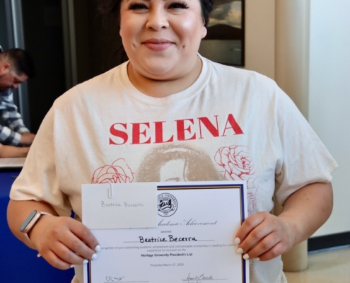 Photograph of a person holding a certificate of achievement from Heritage University, standing indoors near a window and a blue table. The person wears a white Selena t-shirt, and the certificate prominently displays the name Beatriz Becerra along with signatures and the university logo.