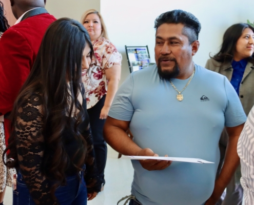 Photograph of a group of people engaged in conversation at an indoor event or gathering. A woman with long dark hair wearing a black lace top is interacting with a man in a light blue shirt holding a document, while other attendees stand in the background.