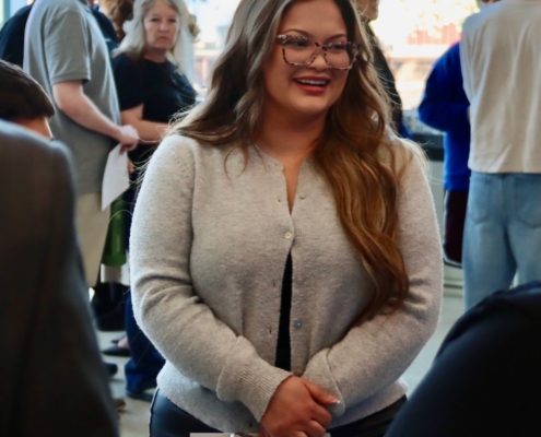 Photograph of a group of people gathered indoors, likely at an event or meeting, with natural light coming through large windows in the background. A woman in the foreground wearing a light gray cardigan holds a smartphone and a piece of paper, while others stand and converse behind her.