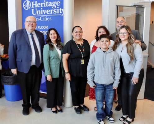 Photograph of a group of seven people standing indoors near a Heritage University Tri-Cities banner, indicating an event or gathering at the university. Individuals are dressed in business casual and casual attire, suggesting a formal yet approachable setting.