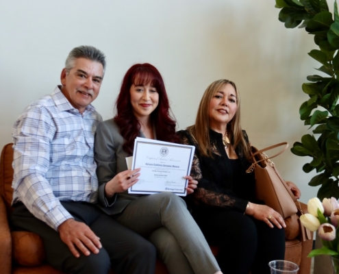 Photograph of three people sitting on a brown couch, with the person in the middle holding a certificate. The setting includes a plant with green leaves and a bouquet of flowers on a table nearby, suggesting a celebratory or formal occasion.