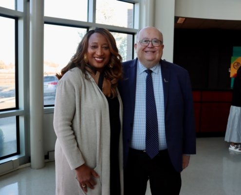 Photograph of two people standing indoors near large windows with natural light, dressed in business casual attire. Background shows other individuals and a sign with partially visible text, suggesting a social or professional event.