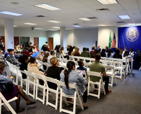 Photograph of a formal indoor event with attendees seated on white folding chairs facing a panel of speakers near flags and a large blue banner. The setting suggests a community meeting or official presentation, with diverse participants attentively listening in a well-lit room.