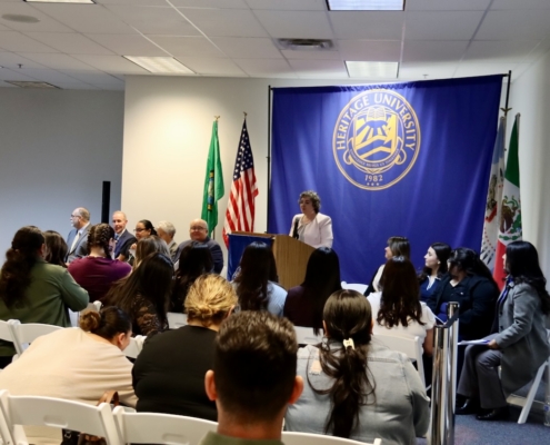Photograph of a formal event at Heritage University featuring a speaker at a podium in front of a blue banner with the university seal and flags of the United States, Washington state, and Mexico. Audience seated in white chairs faces the speaker, with several individuals seated on stage alongside the podium.