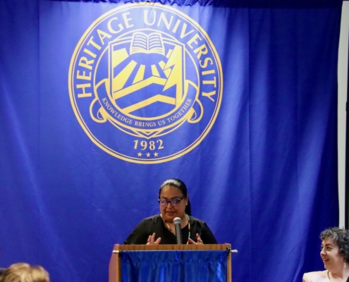 Photograph of a speaker at a podium in front of a large blue banner displaying Heritage University’s gold emblem and motto, "Knowledge Brings Us Together," with the founding year 1982. Audience members are partially visible, indicating a formal event or presentation setting.