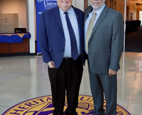 Photograph of two men in business attire standing on a circular Heritage University emblem on a polished floor inside a building lobby. Background includes a Heritage University sign and office furniture, indicating a formal or professional setting at the university.