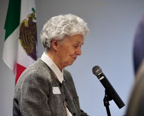 Photograph of a person speaking into a microphone during an event or presentation, with a Mexican flag visible in the background. The person wears a gray textured jacket and white shirt, suggesting a formal or professional setting.