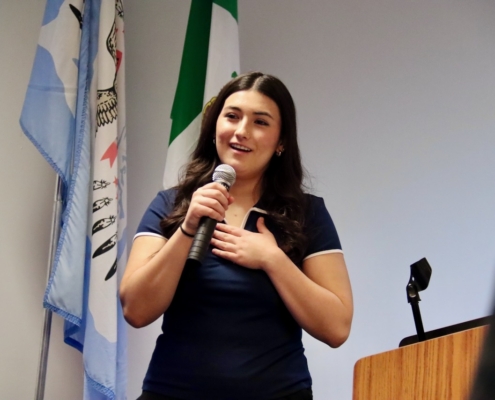 Photograph of a woman speaking into a microphone while standing near a wooden podium, likely during a presentation or speech. Three flags with distinct designs and colors are visible in the background, suggesting a formal or official setting.