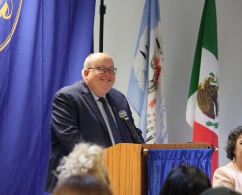 Photograph of a man in a suit speaking at a podium with a microphone during an event or conference. Background includes blue curtain with emblem, and flags of Yakama Nation and Mexico, indicating an international or diplomatic context.