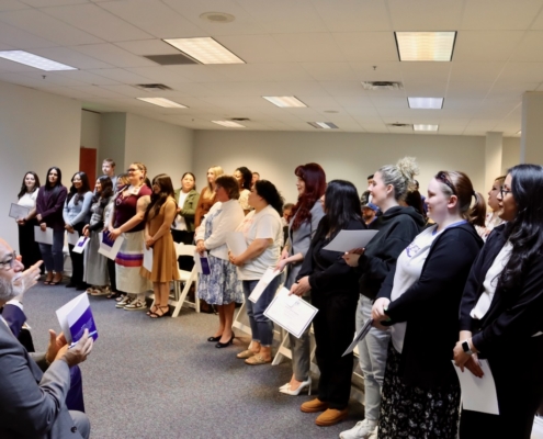 Photograph of a group of people standing in a room, likely attending a formal event or ceremony, with some holding certificates or papers. The setting includes fluorescent ceiling lights and a carpeted floor, with individuals dressed in business casual to formal attire, suggesting a professional or celebratory context.