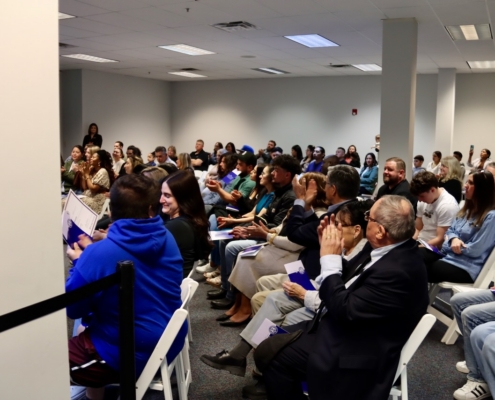 Photograph of a diverse group of people seated in rows inside a conference or seminar room, attentively listening and clapping. The setting includes white folding chairs, neutral-colored walls, and fluorescent ceiling lights, indicating a formal or educational event.