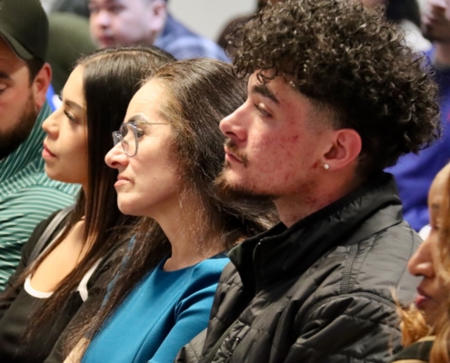 Photograph showing a group of people seated closely together, likely attending an event or presentation. Focus is on individuals wearing casual clothing, with one person in a blue shirt and another in a black jacket, highlighting attentive posture and engagement.