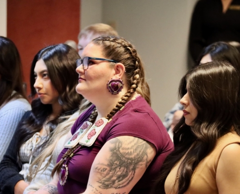 Photograph of a group of people seated indoors, attentively facing forward, likely attending an event or meeting. Notable details include a woman with braided hair, a tattoo on her arm, and large earrings, while others wear casual and formal attire.